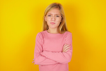 Gloomy dissatisfied caucasian little kid girl wearing long sleeve shirt over yellow background looks with miserable expression at camera from under forehead, makes unhappy grimace