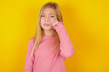 Disappointed dejected caucasian little kid girl wearing long sleeve shirt over yellow background wipes tears stands stressed with gloomy expression. Negative emotion