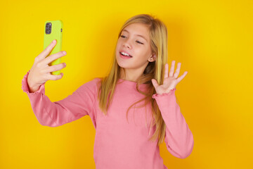 caucasian little kid girl wearing long sleeve shirt over yellow background holds modern mobile phone and makes video call waves palm in hello gesture. People modern technology concept