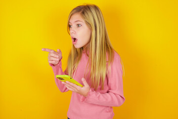 Stunned caucasian little kid girl wearing long sleeve shirt over yellow background points sideways right copy space, recommends product, sees astonishing thing