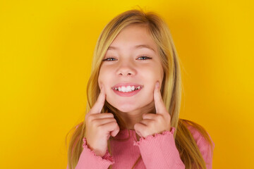 Happy caucasian little kid girl wearing long sleeve shirt over yellow background with toothy smile, keeps index fingers near mouth, fingers pointing and forcing cheerful smile