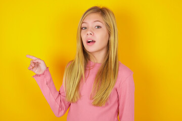 Emotive caucasian little kid girl wearing long sleeve shirt over yellow wall keeps jaw dropped from shock demonstrates amazing promo points right on blank space demonstrates big shopping sale. 