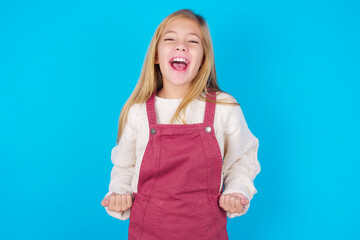 Fototapeta premium caucasian little kid girl wearing jumpsuit over blue background raising fists up screaming with joy being happy to achieve goals.