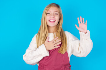 I swear, promise you not regret. Portrait of sincere caucasian little kid girl wearing jumpsuit over blue wall raising one arm and hold hand on heart as give oath, telling truth, want you to believe.