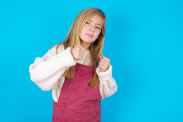 Fototapeta premium Portrait of attractive caucasian little kid girl wearing jumpsuit over blue background holding hands in front of him in boxing position going to fight.
