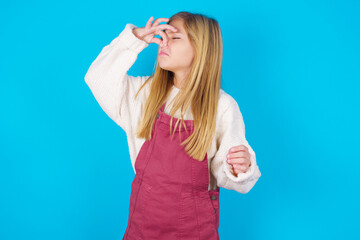 Displeased caucasian little kid girl wearing jumpsuit over blue background plugs nose as smells...