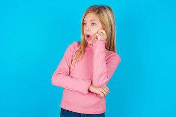Astonished caucasian little kid girl wearing long sleeve shirt over blue background looks aside surprisingly with opened mouth.