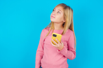 caucasian little kid girl wearing long sleeve shirt over blue background holds mobile phone uses high speed internet and social networks has online communication. Modern technologies concept