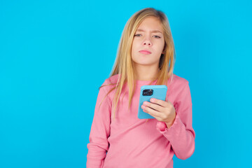 Upset dissatisfied caucasian little kid girl wearing long sleeve shirt over blue background uses mobile software application and surrfs information in internet, holds modern mobile hand