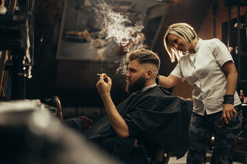 Young bearded man getting haircut by hairdresser and smoking a cigarette