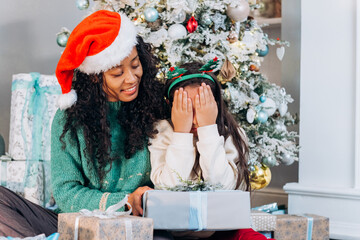 African American mother with curly hair surprises brunette daughter wearing holiday headband with presents smiling near Christmas tree