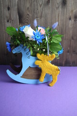 bouquet of flowers in rocking horse vase on wooden background and woolen felted moose toy