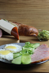 breakfast traditional scrambled eggs fried eggs two ham sausages and cucumber salad on a gray plate on the table. next to it are products for cooking breakfast sausage bread. Vertical photo