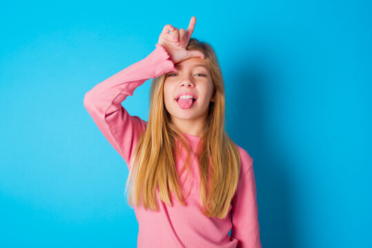 Funny caucasian little kid girl wearing long sleeve shirt over blue background makes loser gesture mocking at someone sticks out tongue making grimace face.