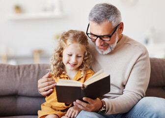 Cute little girl granddaughter reading book with positive senior grandfather