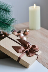 Decorative brown gift box on a wooden table with brown ribbon and brown Christmas balls and Christmas tree with a candle in a background