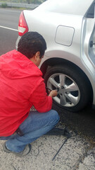 Latino adult man changes the flat tire of his car with tools on the side of the road in the sun
