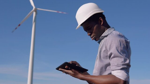 An African-American electrician in a helmet stands against the backdrop of a windmill at an air power plant and looks at a tablet and swipes across the screen. Renewable energy concept