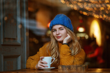 Happy smiling woman holding cup of  coffee or tea, sitting, posing at cafe. Winter vacation, Christmas holidays conception. Model wearing knitted beanie hat, sweater. Copy, empty space for text