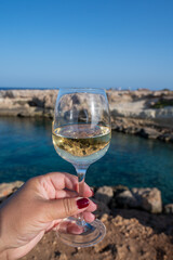 Woman's hand holding glass of white dry white wine with view on rocks and blue sea bay water near Protaras touristic town on Cyprus