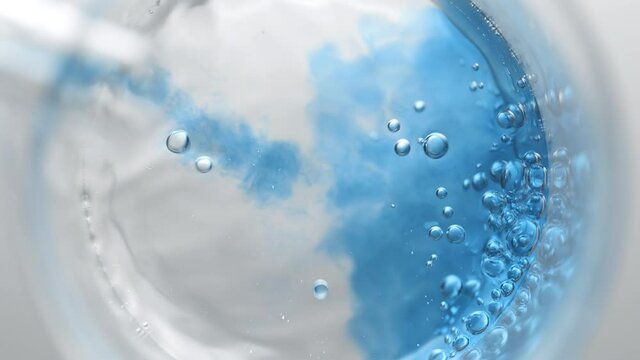 Blue liquid pouring into beaker with water creates different sized bursting bubbles and ripples on its surface on light grey background | Abstract skin nourishing cosmetics mixing concept