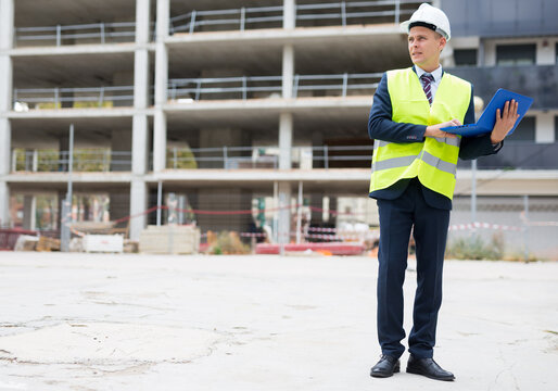 European architect in workwear and white hardhat with laptop in hands standing on building site.