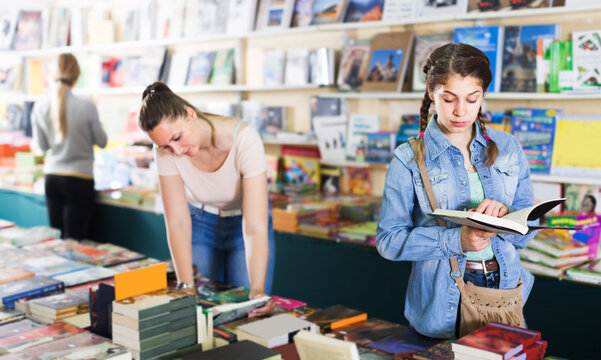 glad woman showing open book to smiling girl in school age in book boutique
