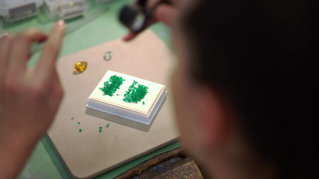 Top view table with precious jewels and blurred female jeweller using tweezers and magnifying glass examining green gems. Unrecognizable Caucasian expert woman checking emerald in workshop indoors