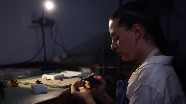 Side view skilled female silversmith polishing ring with manual tool in workshop with blue light. Confident concentrated professional Caucasian woman jeweller working indoors. Slow motion