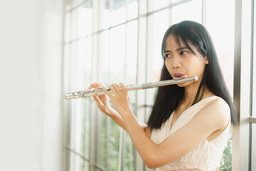 A female flutist performs on her instrument. A young and elegant Asian woman plays the flute - obrazy, fototapety, plakaty