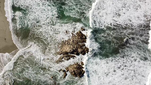 waves break around a small rock in front of the beach of rena majore on sardinia's north-west coast, filmed with a drone from above, sunny weather