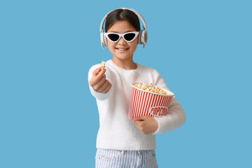 Fototapeta premium Happy cute girl in headphones and sunglasses holding bucket of popcorn on blue background