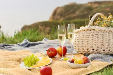 Fotobehang Eetkamer Romantic picnic with sparkling wine, cheese, grapes, apples and peaches on knitted blankets beside a wicker basket in a grassy countryside landscape  © Pixel-Shot