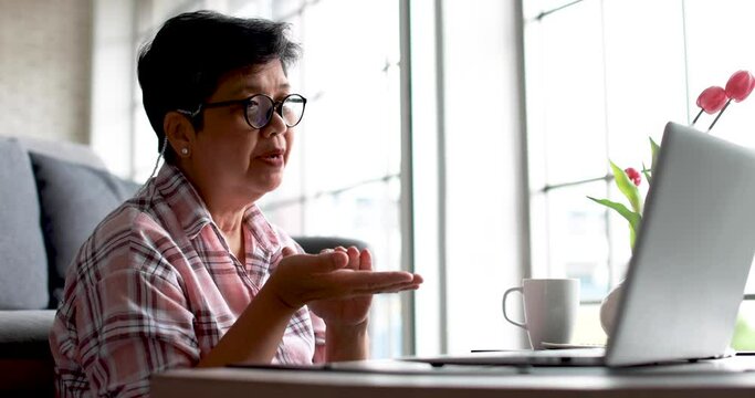Low angle of mature Asian female freelancer gesticulating and explaining data to online colleagues while making video call via laptop in living room at home