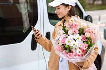 Female courier with bouquet of flowers and phone near car outdoors - obrazy, fototapety, plakaty