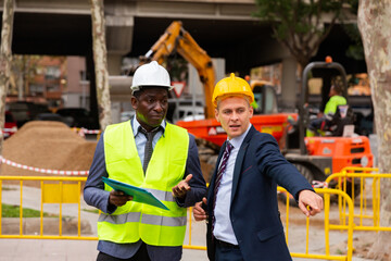 Young confident foreman, controlling the course of laying paving slabs, shows someting to the african-american worker, ..pointing is it