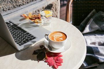 Cup of tasty coffee, autumn leaves and laptop on table in cafe
