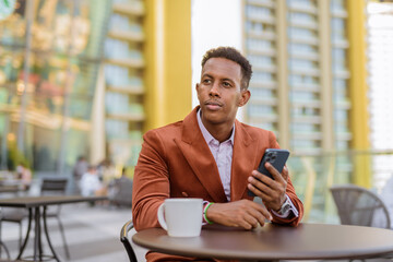 Portrait of handsome black African businessman sitting outdoors in coffee shop using mobile phone