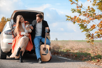 Lovely young couple with cute dog and guitar sitting in car trunk outdoors