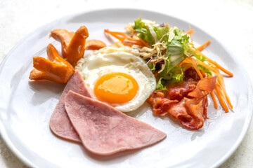 Breakfast menu, fried egg, ham, bacon, sausage, and salad. Breakfast on white plate on white stone table, natural light.