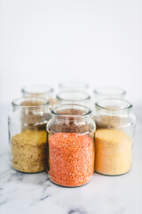 healthy whole grains and legumes in clear pantry jars on marble background including quinoa rice buckwheat lentils and barley, simple ingredients concept