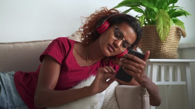 Podcast to inform and entertain audience. Relaxed young African-American teenage lady with modern earphones spbas and cellphone enjoys music on sofa at home