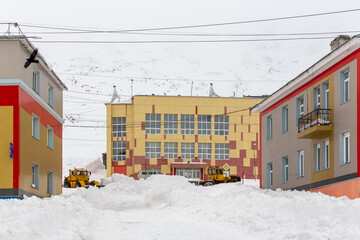 Snow-covered street in a northern town in the Arctic. Bulldozers remove snow. Clearing snow from streets after a blizzard. Large snowdrifts near buildings. Provideniya, Chukotka, Far North of Russia.