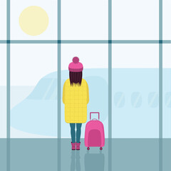 A girl in a bright yellow down jacket and a pink hat with a pink suitcase. A girl in the airport building , looking at the plane through the glass.