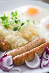 Pork sausages with onions, eggs, mashed potatoes and microgreens. Hearty breakfast. Dark wooden background, white ceramic plate. Top view.