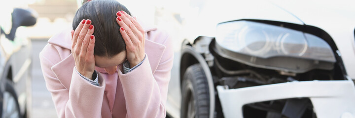 Upset young woman sits with head bowed next to wrecked car