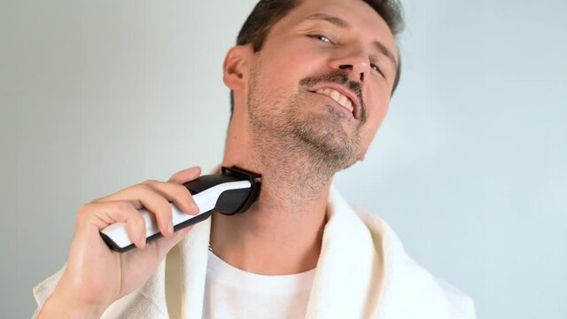 Handsome young unshaven man with towel on shoulders shaving his neck area with modern electric razor trimer while looking at camera, indoors. Daily routine male self-care