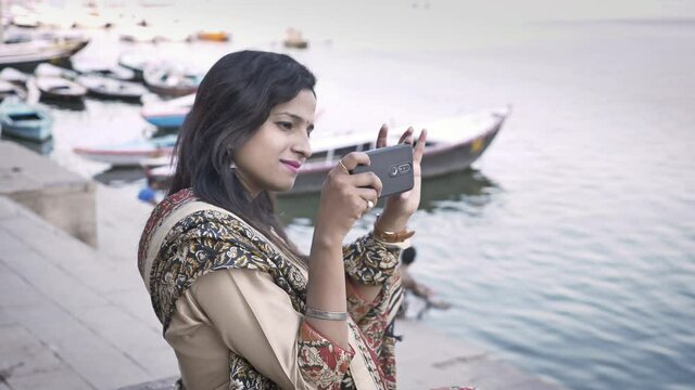 A young beautiful woman in traditional Salwar Kameez and wearing spectacles is taking images or photos with her cellphone camera near a riverbank.