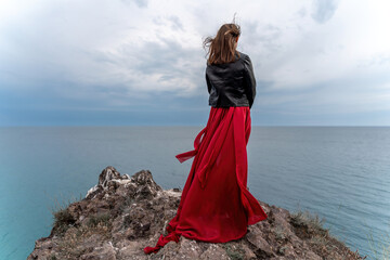 A woman in a red dress and leather jacket stands above a stormy sky, her dress fluttering, the fabric flying in the wind.