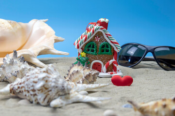 A small Christmas toy in the shape of a gingerbread house stands on a sandy beach next to beautiful shells. Merry Christmas by the warm sea.